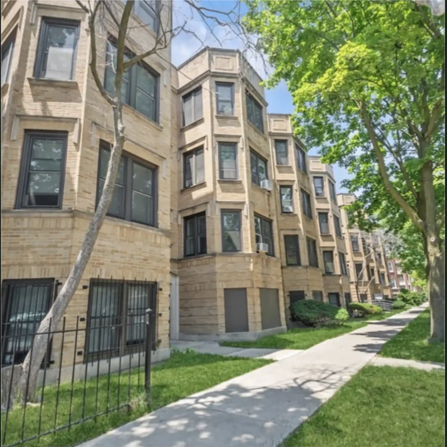 A row of multi-story brick apartment buildings with bay windows, black window frames, and a tree-lined sidewalk on a sunny day.