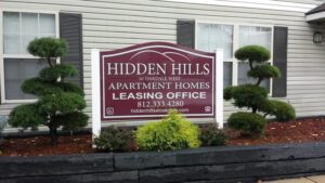Sign for Hidden Hills Apartment Homes Leasing Office is displayed in front of a building with trimmed bushes and landscaped mulch. Contact number and website are also shown on the sign.