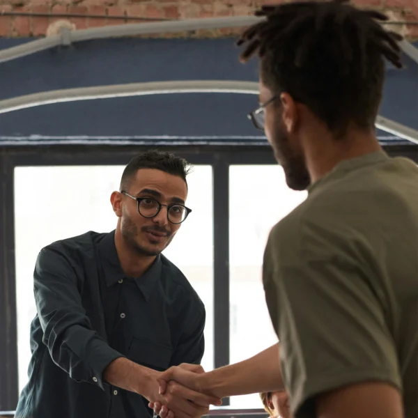 Two men are shaking hands indoors, one facing the camera and the other with his back to the camera, in front of large windows.