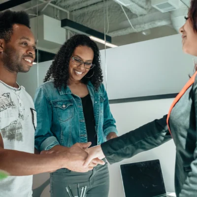 Two people smile while shaking hands with a third person in an office setting, with a laptop and office supplies visible on the desk.