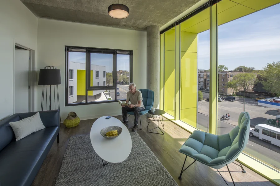 A man sits reading in a modern, brightly lit lounge with large yellow-framed windows, contemporary furniture, and a view of buildings and trees outside.