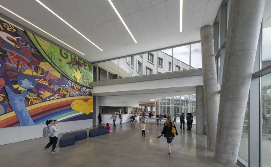Spacious modern lobby with large, colorful mural on the wall, tall windows, concrete columns, and people walking or standing throughout the area.