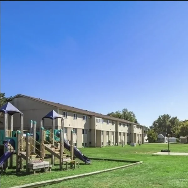 A playground with slides and climbing structures sits on a grassy area next to a row of beige apartment buildings under a clear blue sky.
