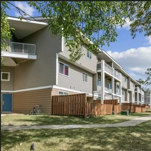 Three-story apartment building with balconies, beige siding, and a wooden fence along the ground floor. A bicycle is parked near the entrance, and trees and grass line the sidewalk.