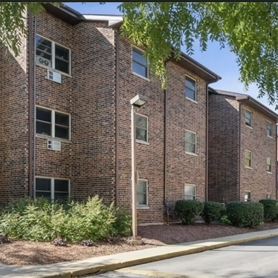 A three-story brick apartment building with multiple windows, window air conditioning units, shrubs, and a sidewalk in front.