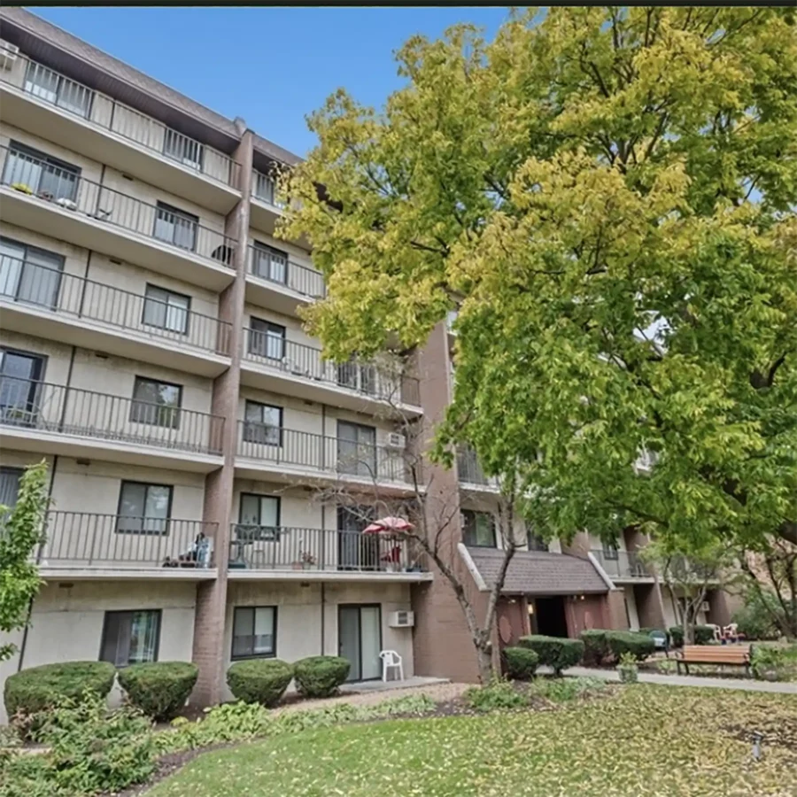Four-story apartment building with balconies, surrounded by trimmed bushes, a large tree with green leaves, and a grassy courtyard under a clear blue sky.