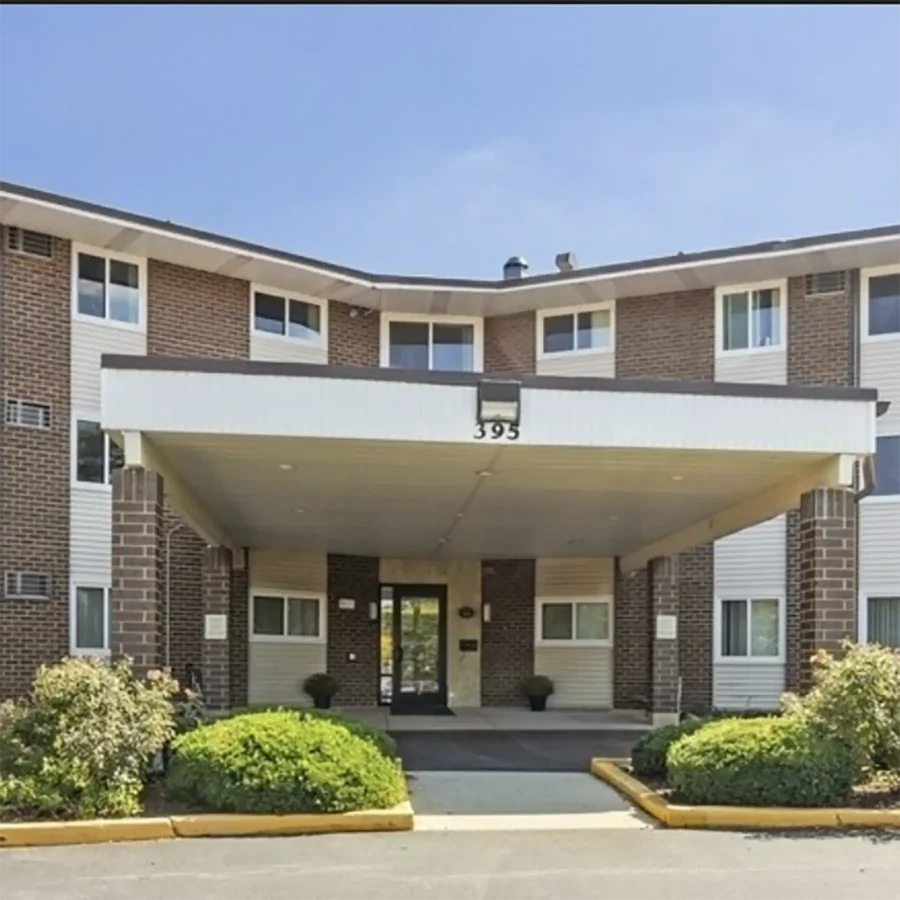 Front entrance of a three-story brick and siding apartment building with a covered driveway and landscaped bushes.