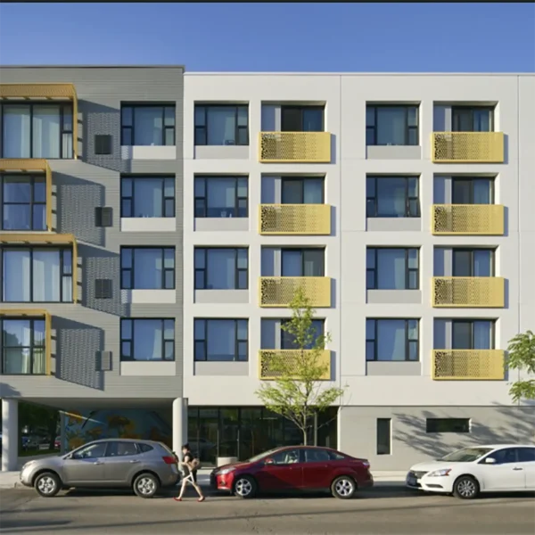 A modern four-story apartment building with yellow balcony railings, large windows, and parked cars in front; a person walks on the sidewalk.