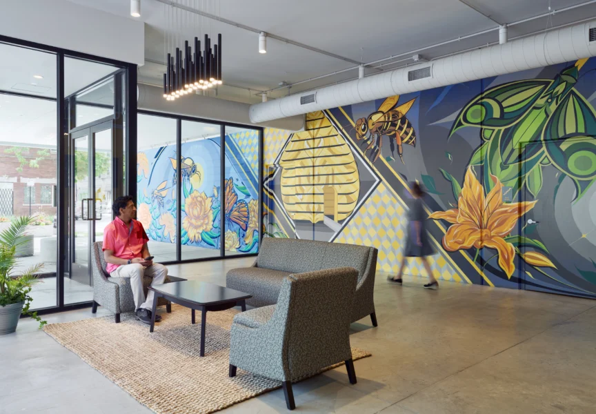 A man sits on a sofa in a modern lobby with large, colorful murals of insects and flowers on the walls; another person walks by in the background.