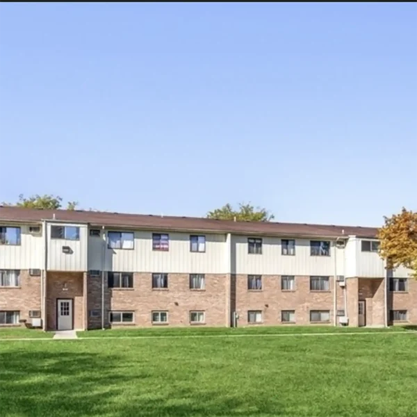 A two-story apartment building with a brick and light-colored exterior, multiple windows, and a large grassy lawn in front under a clear sky.