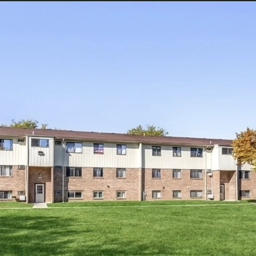 A two-story apartment building with a brick and light-colored exterior, multiple windows, and a large grassy lawn in front under a clear sky.