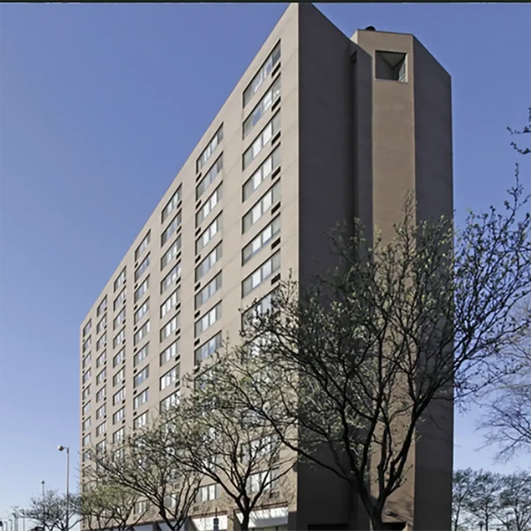 A multi-story rectangular apartment building with uniform windows, brown exterior, and leafing trees lining the sidewalk in front, under a clear blue sky.