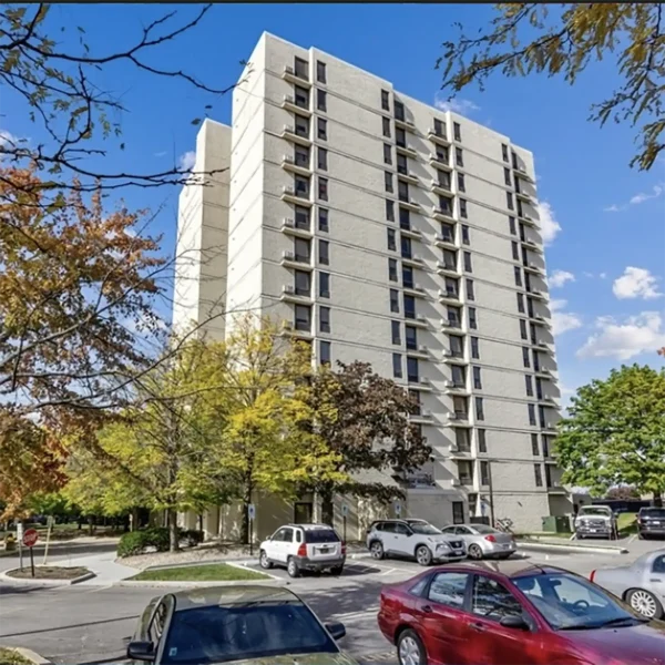 A tall, light-colored apartment building with balconies is surrounded by trees, cars, and a parking lot under a partly cloudy sky.