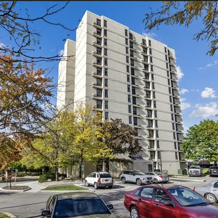 A tall, light-colored apartment building with balconies is surrounded by trees, cars, and a parking lot under a partly cloudy sky.