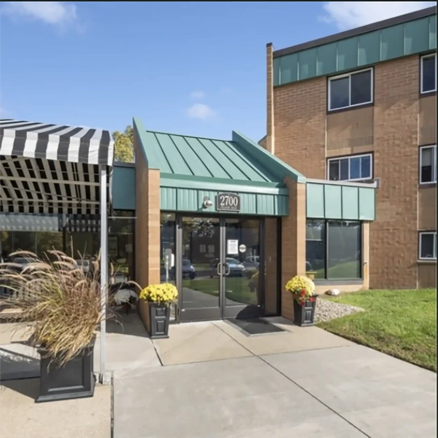 Entrance to a brick building with green accents, glass doors, two potted yellow flowers, and a striped awning on the left side.