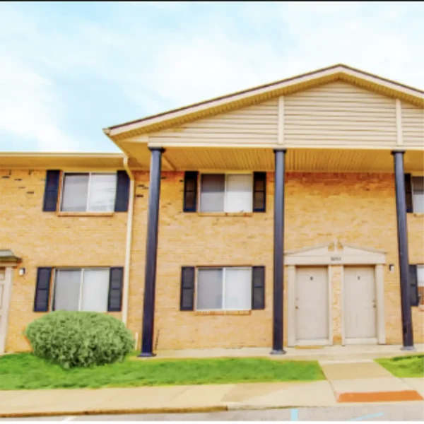 Two-story brick apartment building with four black columns, black shutters, two front doors, and a small shrub on a grassy area.