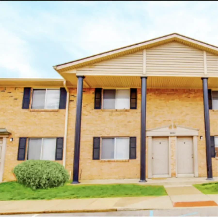 Two-story brick apartment building with four black columns, black shutters, two front doors, and a small shrub on a grassy area.