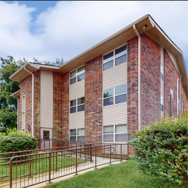 A three-story brick and siding apartment building with a sloped roof, multiple windows, and a metal accessibility ramp leading to the entrance.