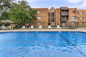 Outdoor swimming pool with lounge chairs, tables, and umbrellas in front of a three-story brick apartment building.
