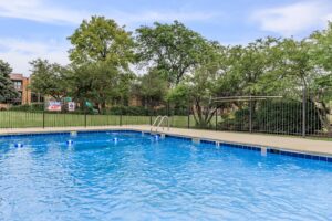 Outdoor swimming pool with clear blue water, surrounded by a safety fence and greenery, under a partly cloudy sky.