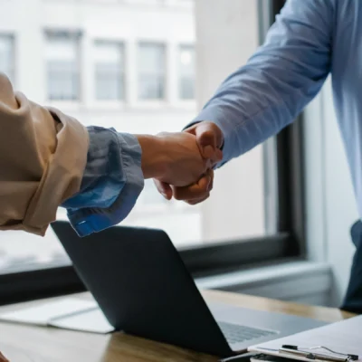 Two people in business attire shake hands across a desk with a laptop, notepad, and pen, in an office setting with large windows.
