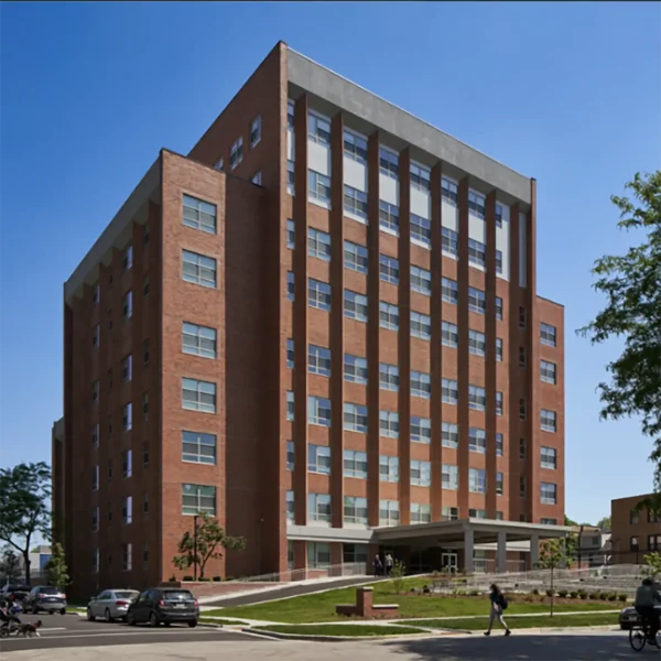 A multi-story red brick building with large windows, set on a landscaped lot with parked cars and a few people walking nearby under a clear sky.