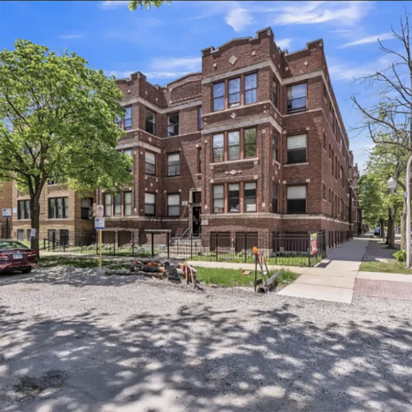A three-story red brick apartment building with multiple windows, located on a corner lot with trees, a sidewalk, and a partially gravel-covered street in front.