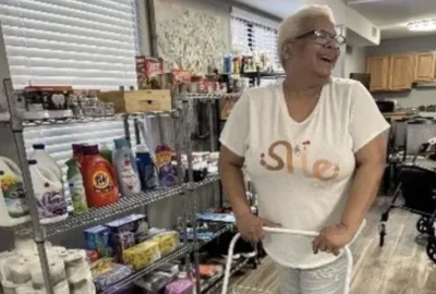 A woman with short blond hair stands smiling in a room with shelves stocked with household supplies such as detergent, paper towels, and cleaning products.