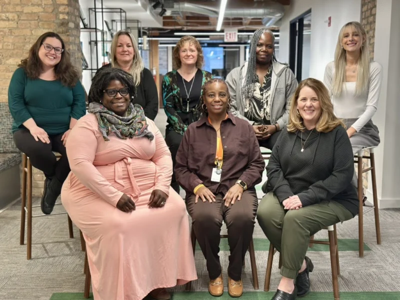 Eight women pose for a group photo indoors, with four seated in front and four standing behind, all smiling at the camera.