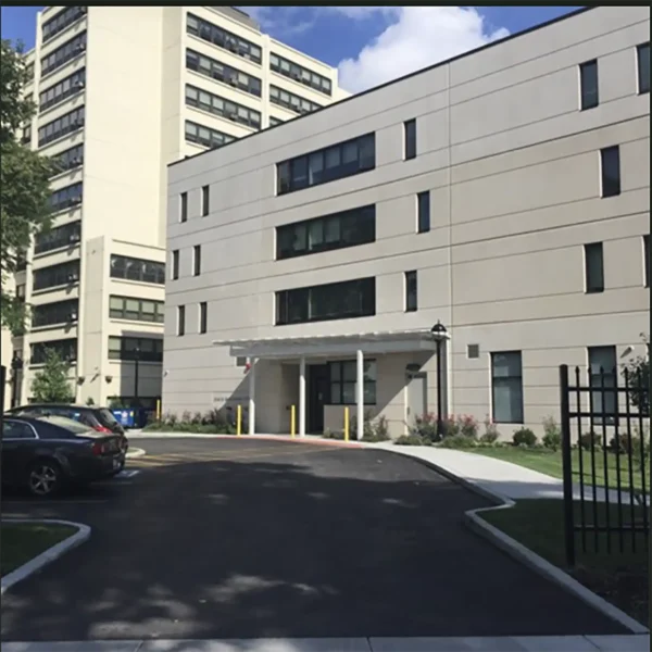 A modern, multi-story beige building with large windows and a driveway, surrounded by parked cars and a black metal fence.