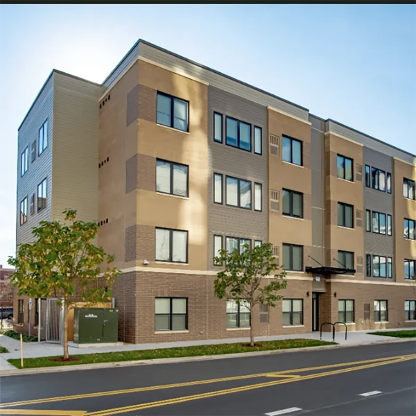 A modern three-story apartment building with tan and brown brick facade, large windows, and small trees along a paved street.