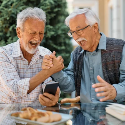 Two older men sit outdoors at a table, smiling and arm wrestling, with food and a smartphone in front of them.