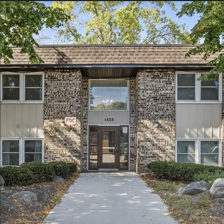 A two-story apartment building with brick and siding exterior, labeled "1428" above the entrance, surrounded by bushes and trees.