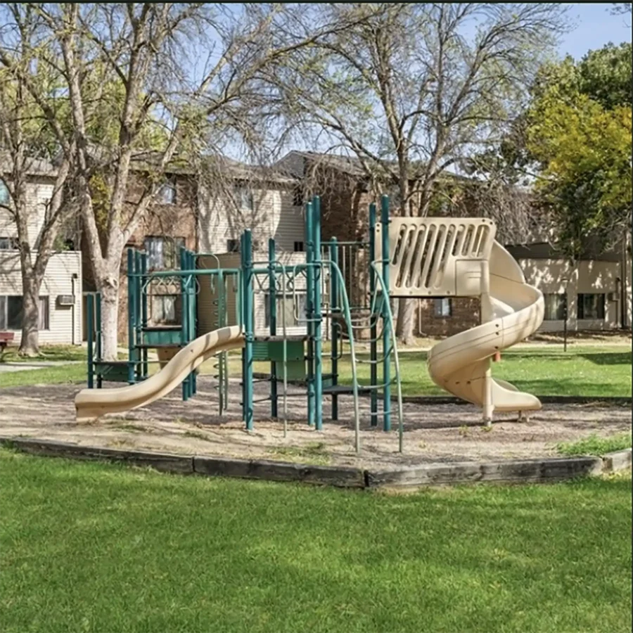 Outdoor playground with slides, climbing structures, and bridges on a grassy area, surrounded by trees and apartment buildings in the background.