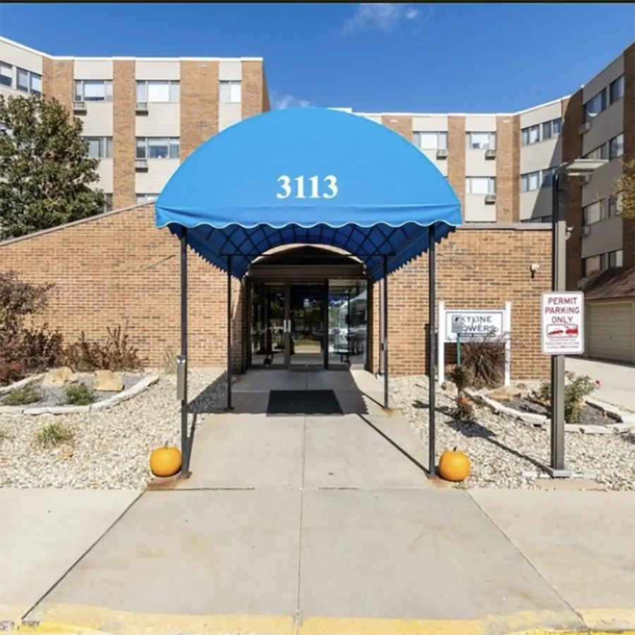 Entrance to a brick apartment building with a blue awning labeled "3113," flanked by two pumpkins and parking signs, on a clear day.