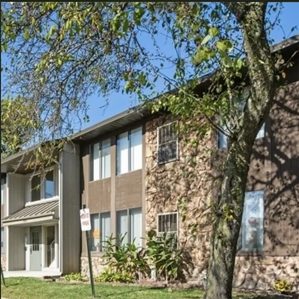 Two-story apartment building with brown siding and stone accents, partially shaded by a tree, with a lawn and a sidewalk in front.