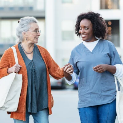 An elderly woman walks outdoors with a caregiver wearing scrubs. They are holding hands and both carrying tote bags.
