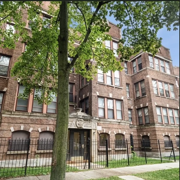 A four-story brick apartment building with large windows, black metal fencing, and a tree in front, under a clear blue sky.