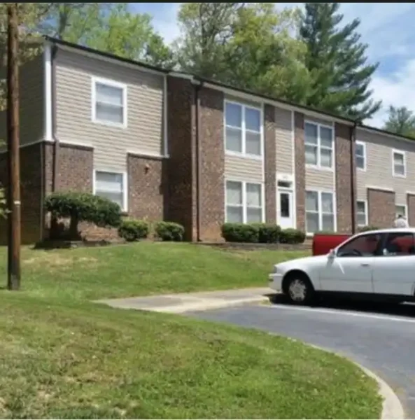 A two-story apartment building with brick and beige siding, surrounded by grass and trees, with a white car parked in front.