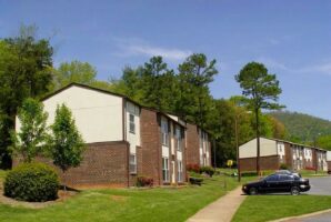 Three two-story brick apartment buildings with white upper walls are surrounded by trees and grass, with a black car parked along the curb on a sunny day.