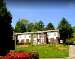 A two-story brick and white apartment building stands on a grassy lawn with shrubs and trees under a clear blue sky.