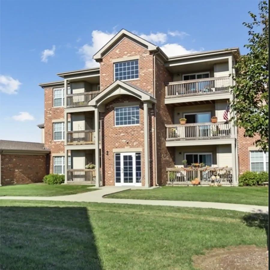 Three-story brick apartment building with balconies, white trim, and double glass doors, surrounded by grass and a few shrubs under a blue sky with scattered clouds.