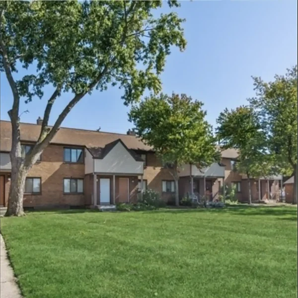 Two-story brick apartment building with multiple entrances, small porches, and a well-maintained lawn with several trees in front.