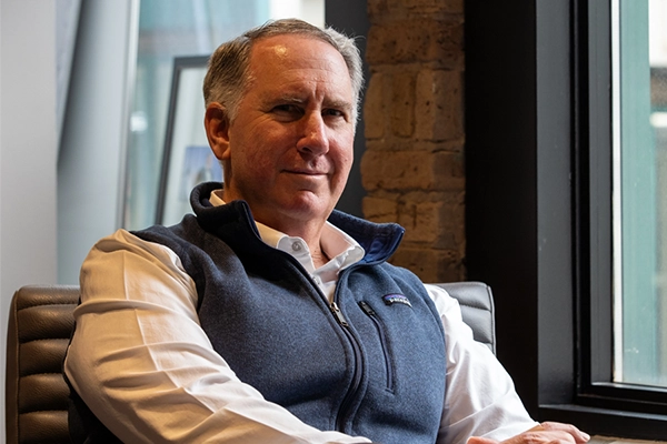 A man in a white shirt and blue vest sits in an office chair next to a window, looking at the camera with a neutral expression.