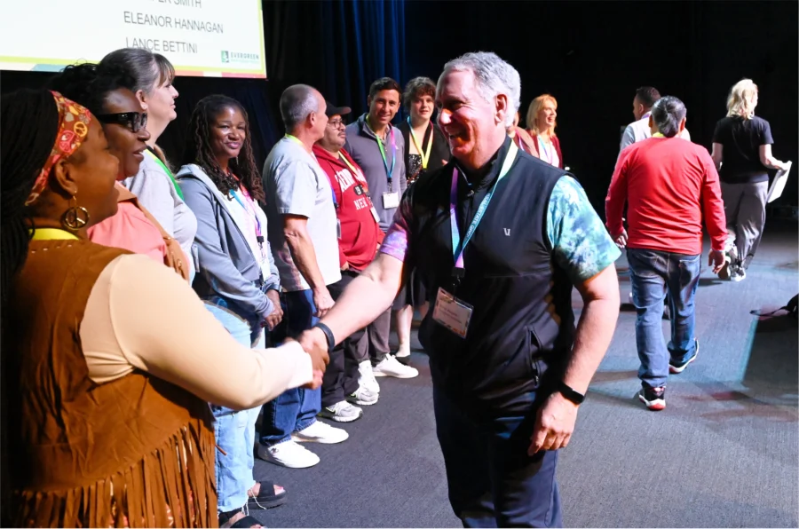 A man wearing a vest smiles and shakes hands with a woman in a line of people on a stage during an event. A presentation screen is visible in the background.