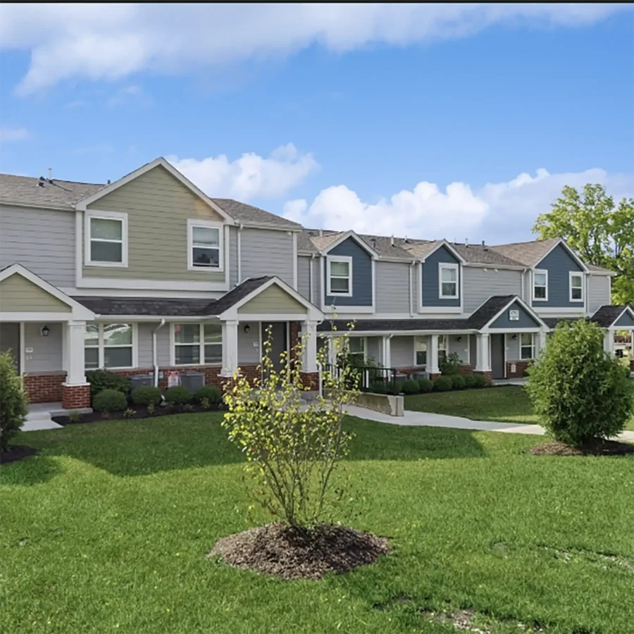 Row of modern two-story townhouses with siding exteriors, small covered porches, and landscaped lawns under a blue sky with scattered clouds.