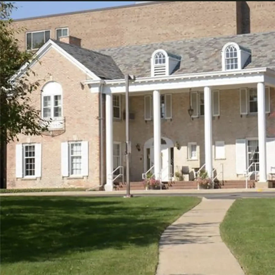 A two-story brick house with white shutters and columns, featuring a covered porch, set on a lawn with a curved sidewalk.