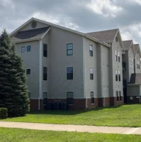 A three-story beige apartment building with red brick trim at the base, multiple windows, and a sloped roof, situated next to a grassy lawn and a pine tree under a cloudy sky.