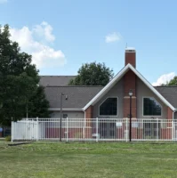 A red brick building with large triangular windows, a white fence, and a chimney, surrounded by green grass and trees under a blue sky.