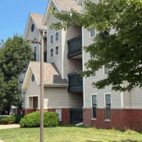 Three-story apartment building with beige siding, red brick base, green balconies, and trees in front, on a sunny day.